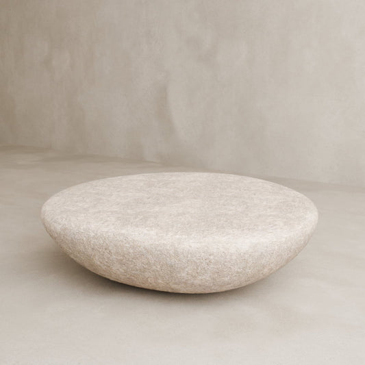 The Pluto Coffee Table, with its round white form and minimalist design, rests on a neutral floor by a textured wall, serving as a chic centerpiece that elevates the room’s modern aesthetic.
