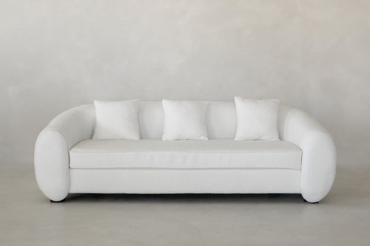 The Polar Bear Sofa is a modern white set with rounded arms and four matching cushions, adding contemporary luxury to any space with a light gray textured wall and floor.