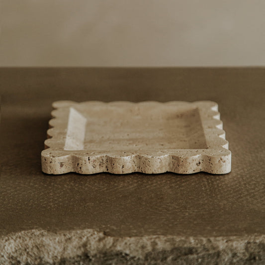 The Remy Tray, a square travertine piece with a modern design and wavy, scalloped edges, rests on a textured brown surface with a neutral background.