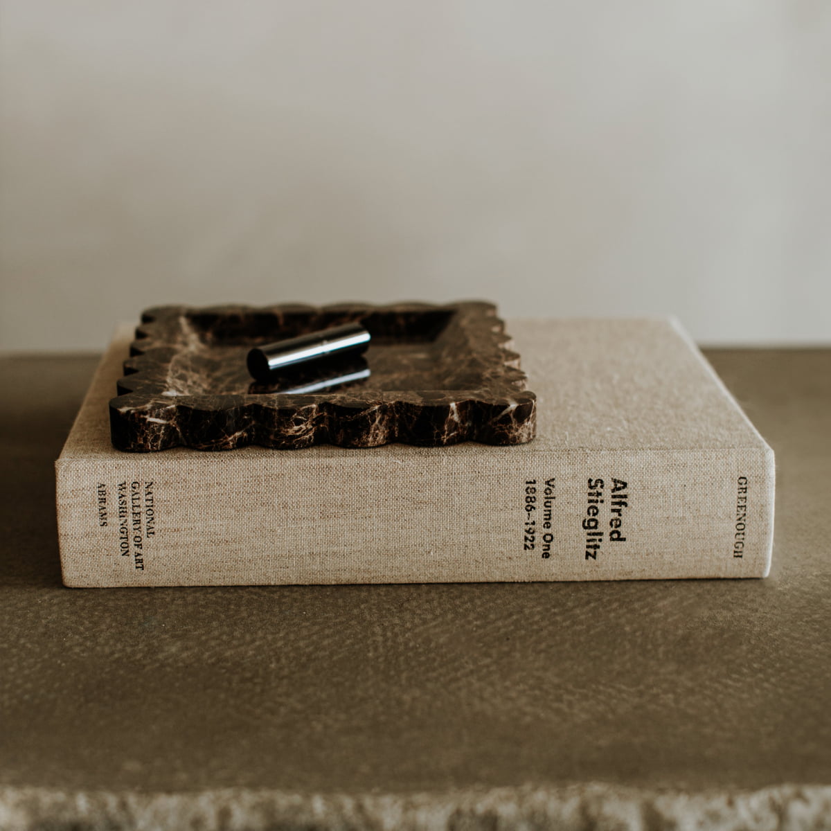 A beige hardcover book labeled Altered Selig 112 and Gestalten sits closed on a taupe surface, topped with the Remy Tray In Marble and a silver cylinder, infusing the scene with modern style.