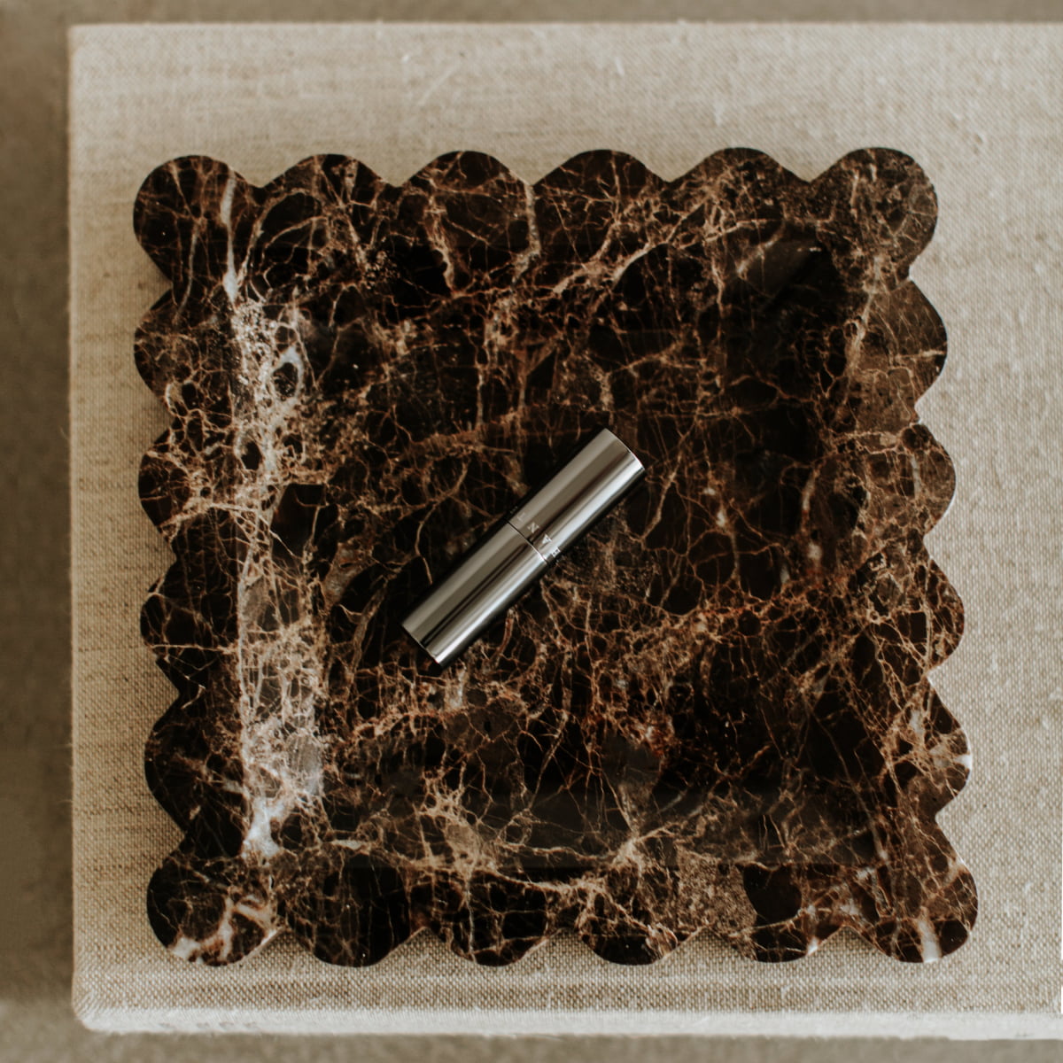 The Remy Tray in Marble, featuring scalloped edges and rich dark brown tones, displays a silver lipstick tube and brings modern elegance to a textured beige linen surface.