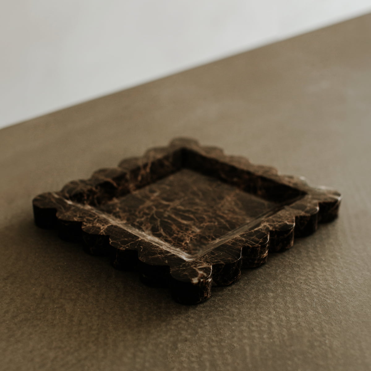 The Remy Tray In Marble—a square, dark marble tray with scalloped edges—adds modern elegance to a textured brown surface, beautifully highlighted by a blurred, light-toned background.