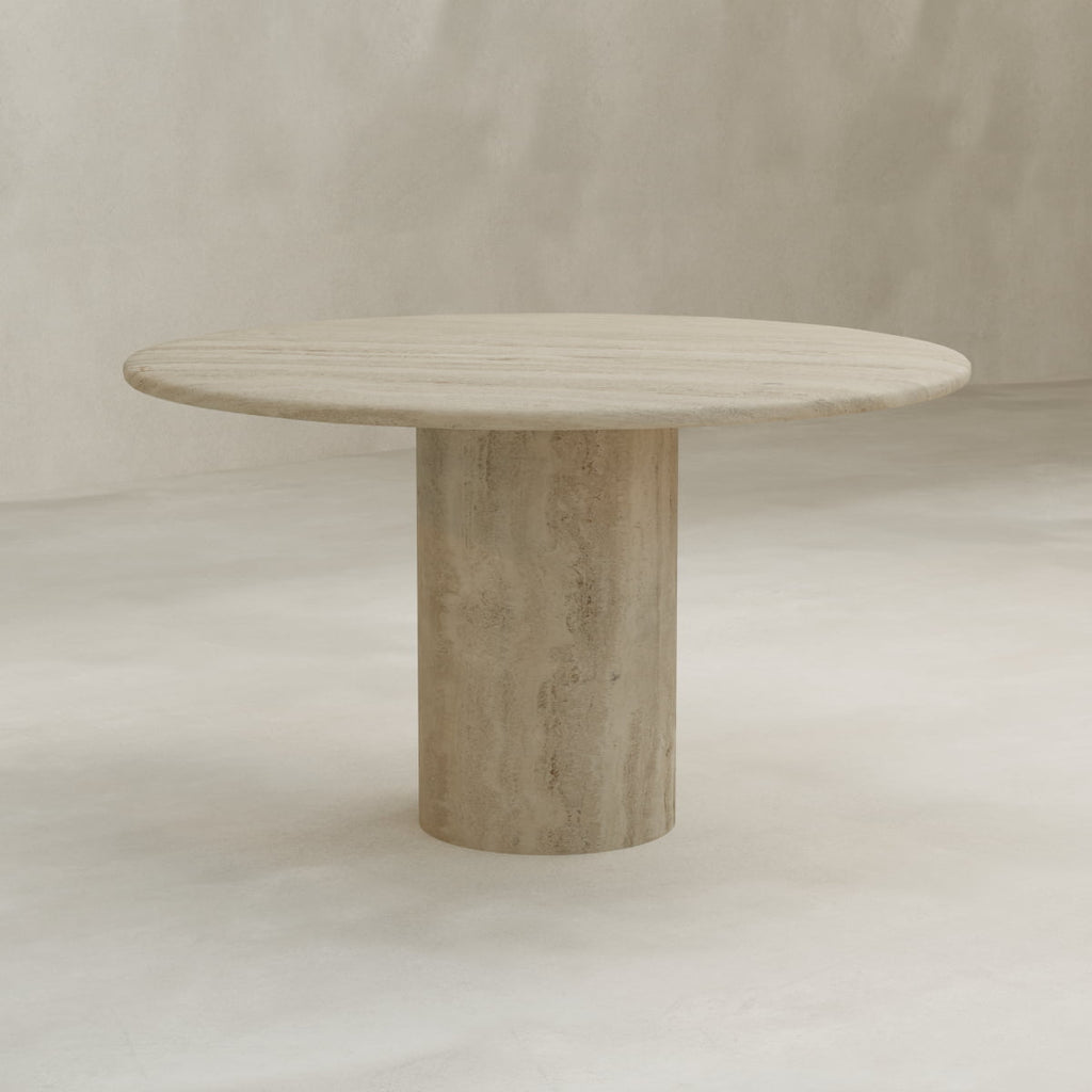 The Renè Travertine Dining Table features a round, beige-colored travertine top and a thick cylindrical base, showcased in a neutral, minimalist setting.