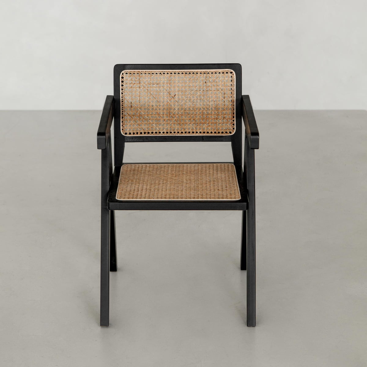 The Rivini Arm Chair features a black wooden frame with hand-woven light cane seat and backrest, shown on a light gray floor against a plain light gray wall.