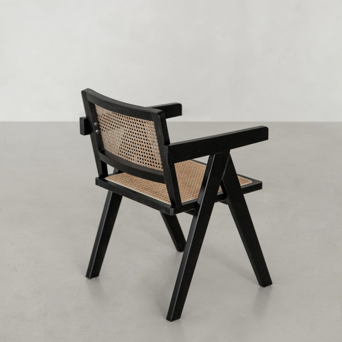 The Rivini Arm Chair, featuring a black wooden frame, angled legs, and hand-woven cane seat and backrest, is placed on a light gray floor against a plain light-colored wall.