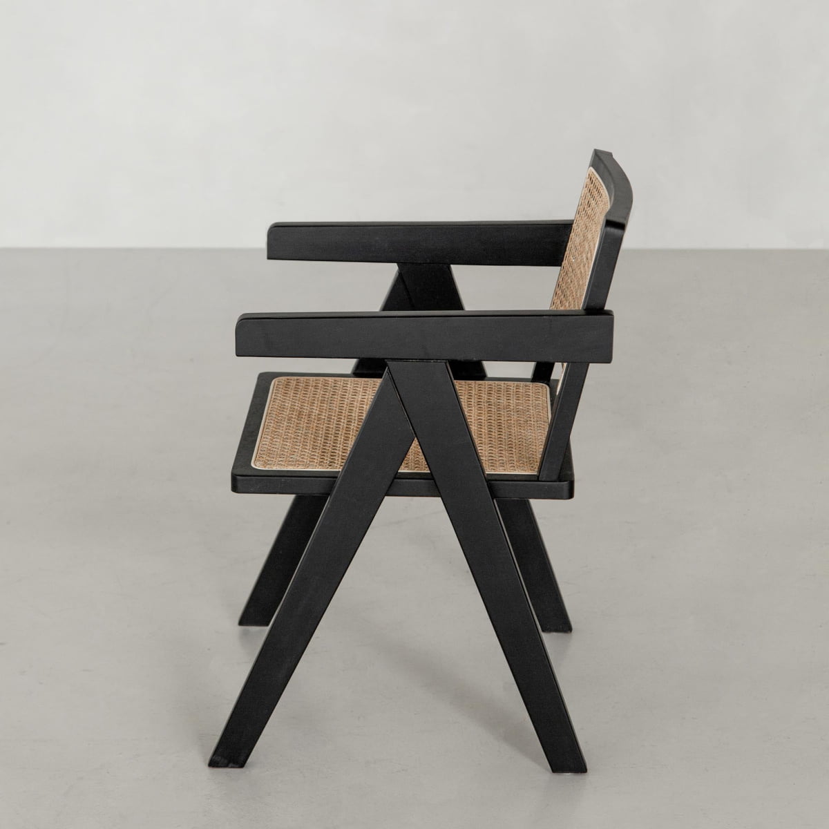 Side view of the Rivini Arm Chair featuring a black wooden frame, angled legs, and rattan weaving on the seat and backrest, set on a light gray floor with a plain light background.