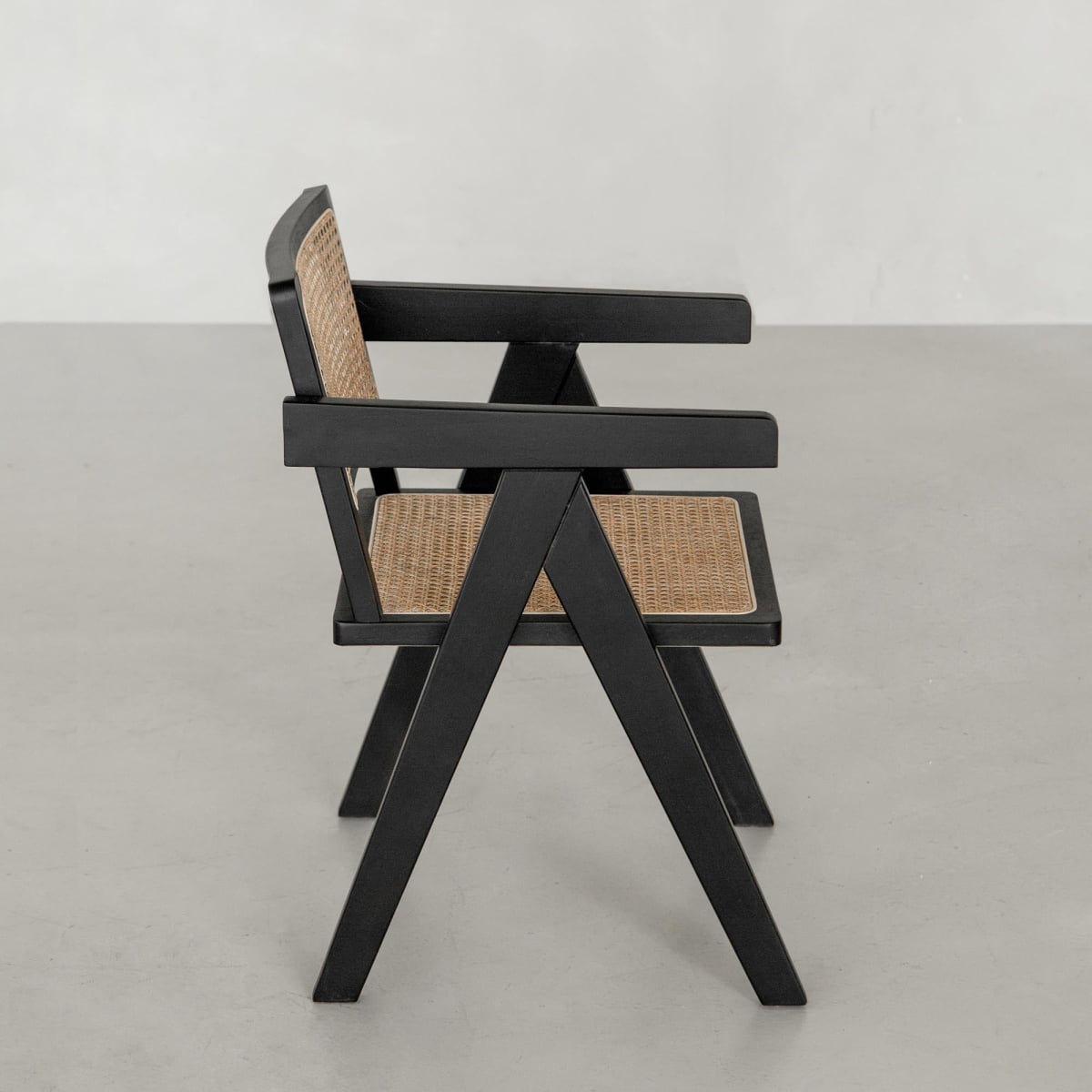 The Rivini Arm Chair features a modern black wooden frame with outward-angled legs and hand-woven cane seat and backrest, shown on a light grey floor against a pale wall.