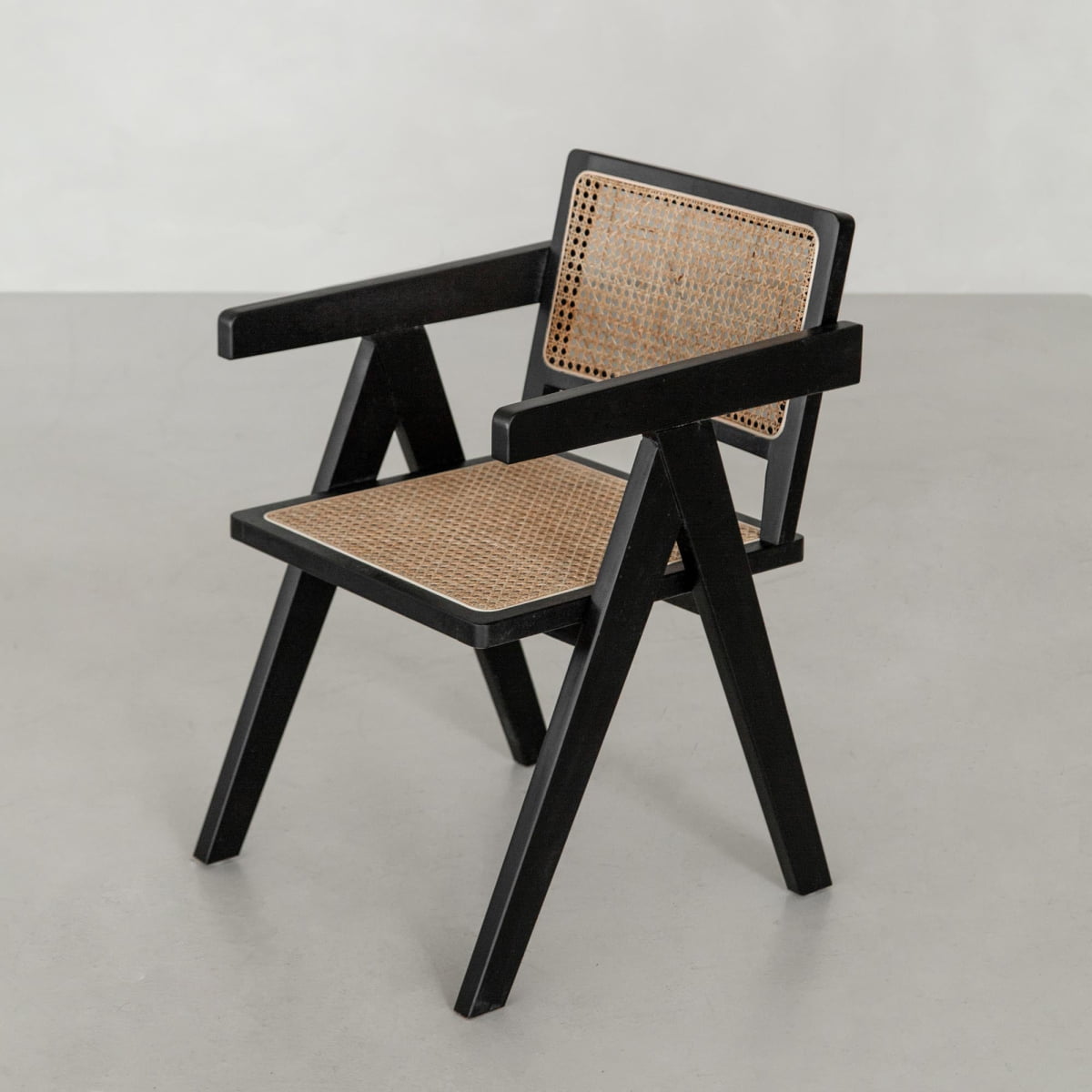 The Rivini Arm Chair is a modern hand-woven cane dining chair with a black wooden frame, angular legs, armrests, and rattan weaving on the seat and backrest, set against a light gray floor and plain light background.