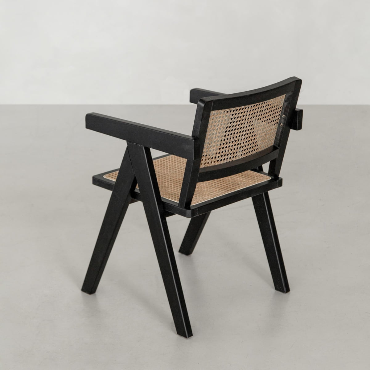 The Rivini Arm Chair is a modern wooden chair with a black frame and angled legs, featuring hand-woven cane details on the seat and backrest, shown on a light gray floor against a plain white background.