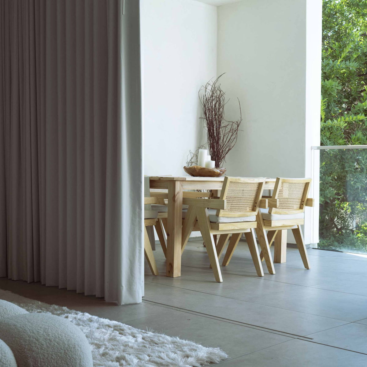 A minimalist dining area features a wooden table, four Rivini Arm Chairs with hand-woven cane backs, a tall branch centerpiece in a vase, and large windows bathed in natural light. A white curtain and rug are partially visible in the foreground.