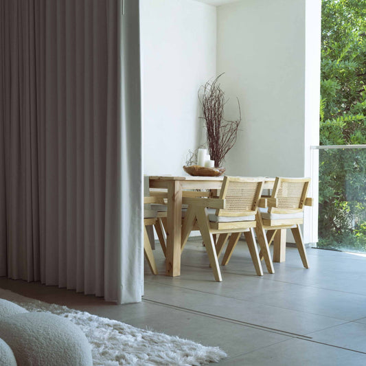 A minimalist dining area features a wooden table, four Rivini Arm Chairs with hand-woven cane backs, a tall branch centerpiece in a vase, and large windows bathed in natural light. A white curtain and rug are partially visible in the foreground.