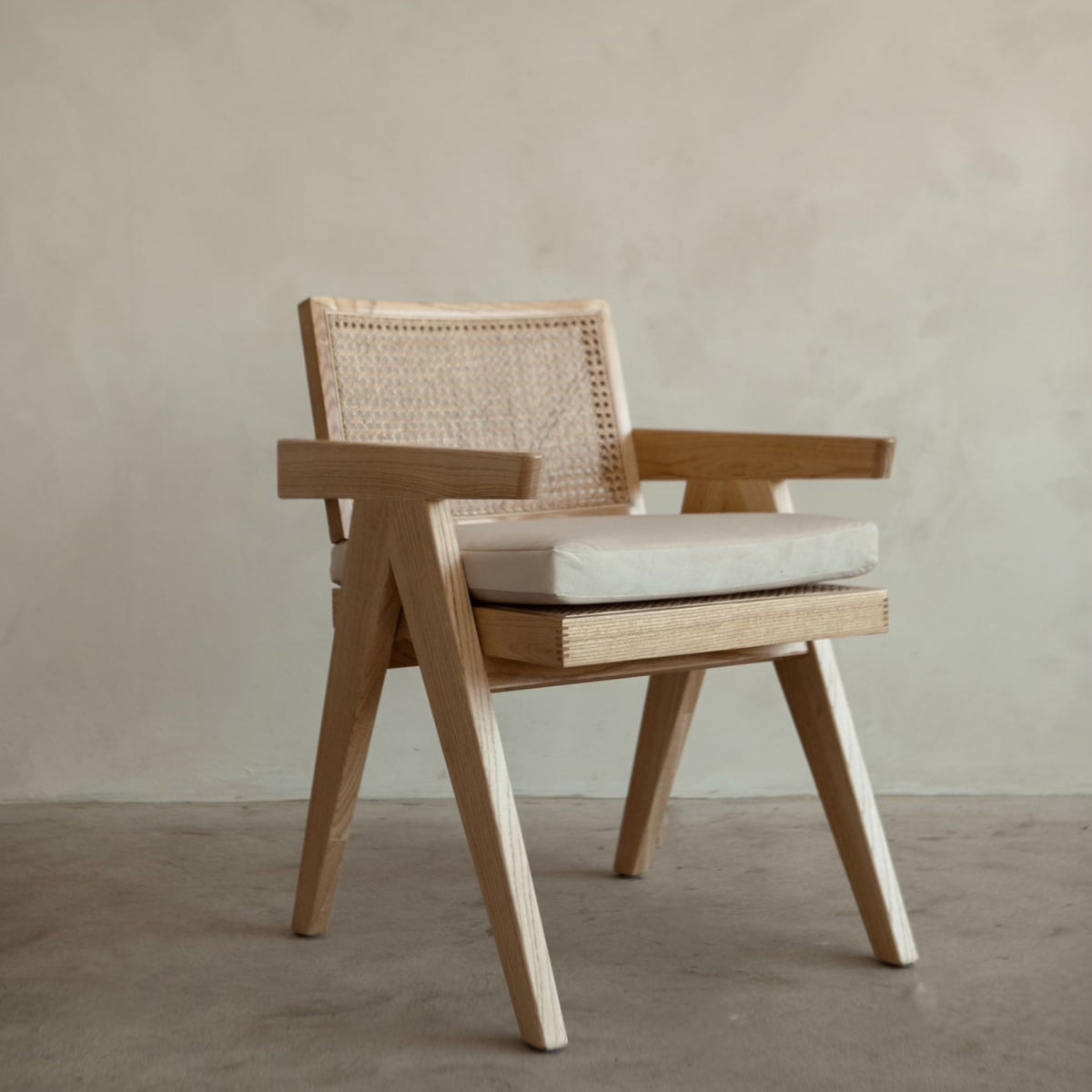 The Rivini Arm Chair features angled wooden legs, a hand-woven rattan cane backrest, and a light beige upholstered seat, shown on a concrete floor against a plain light wall.