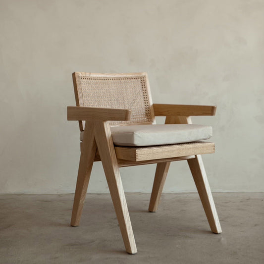 The Rivini Arm Chair features angled wooden legs, a hand-woven rattan cane backrest, and a light beige upholstered seat, shown on a concrete floor against a plain light wall.