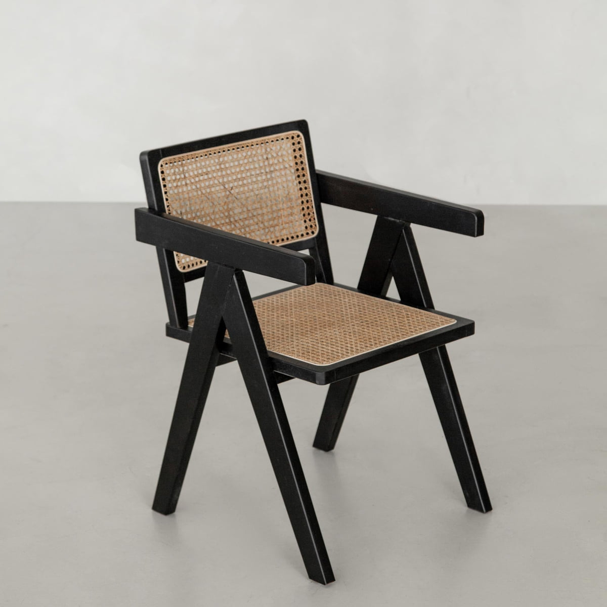 The Rivini Arm Chair features a black angular wooden frame with rattan weaving on the seat and backrest, displayed on a smooth light gray floor against a plain light-colored wall.