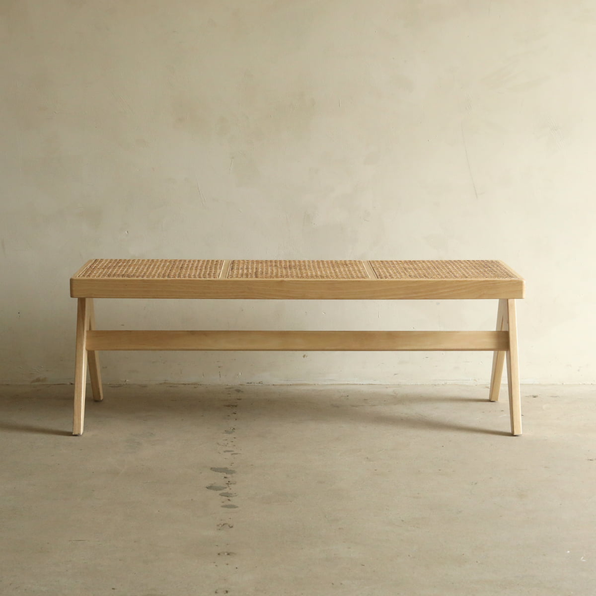 The Rivini Bench, featuring cane detailing and a woven rattan seat, sits against a light wall on a concrete floor. Its minimalist design and natural wood finish create an understated look.