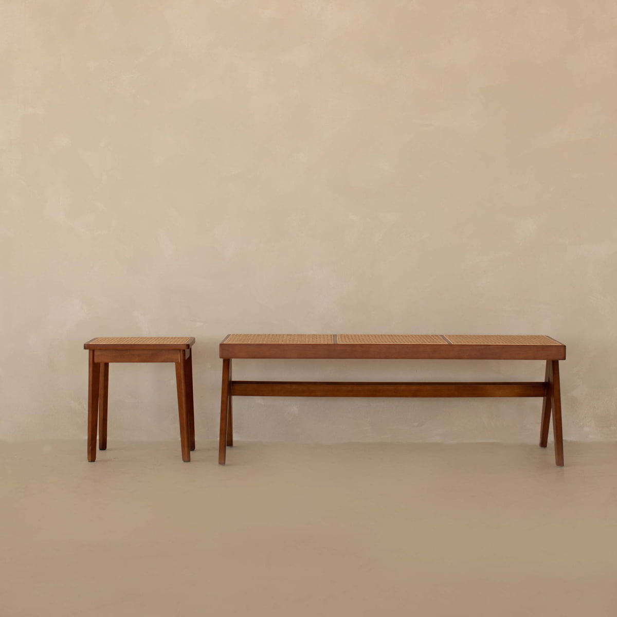 The Rivini Bench Set, featuring a small teak wood stool and matching cane bench with woven seats in Scandinavian style, sits side by side against a plain beige wall.
