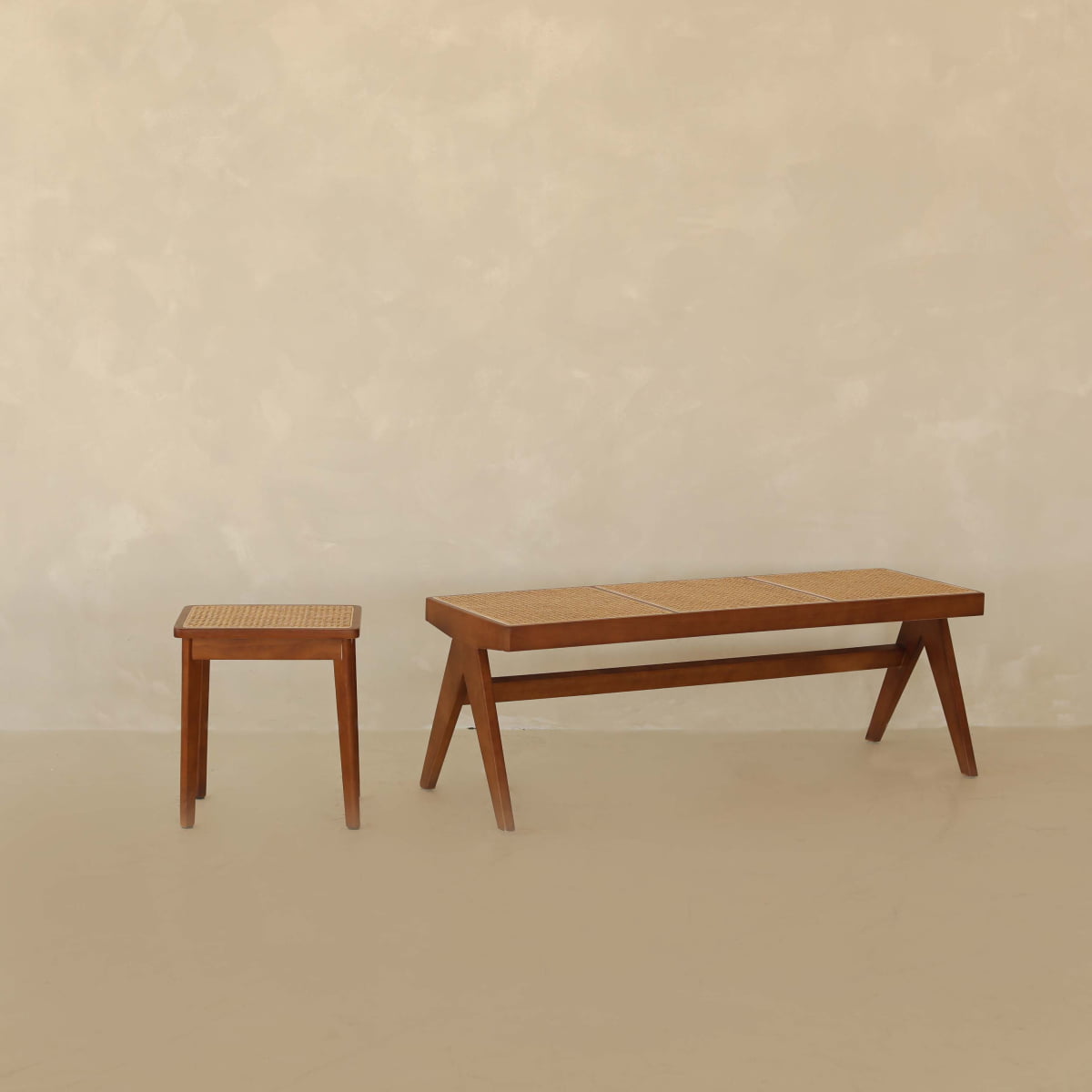 The Rivini Bench Set, with its teak wood stool and matching cane bench featuring woven seats, is displayed in a minimalist setting of beige textured walls and floors, reflecting understated Scandinavian design.