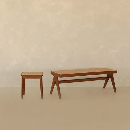 The Rivini Bench Set, with its teak wood stool and matching cane bench featuring woven seats, is displayed in a minimalist setting of beige textured walls and floors, reflecting understated Scandinavian design.