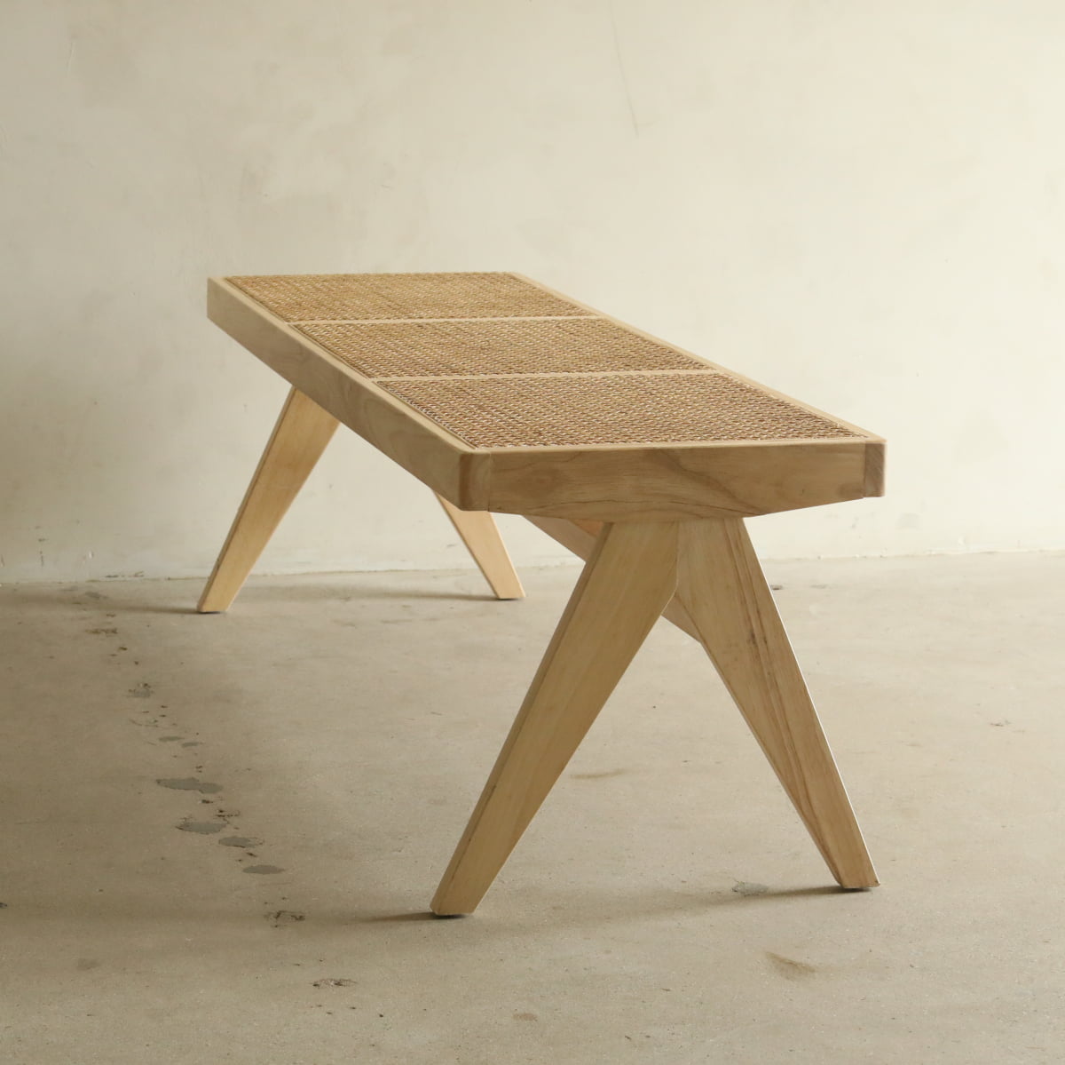 The Rivini Bench, featuring angled wooden legs and woven cane detailing, stands on a plain concrete floor against a neutral wall, bringing Scandinavian-inspired style to the space.