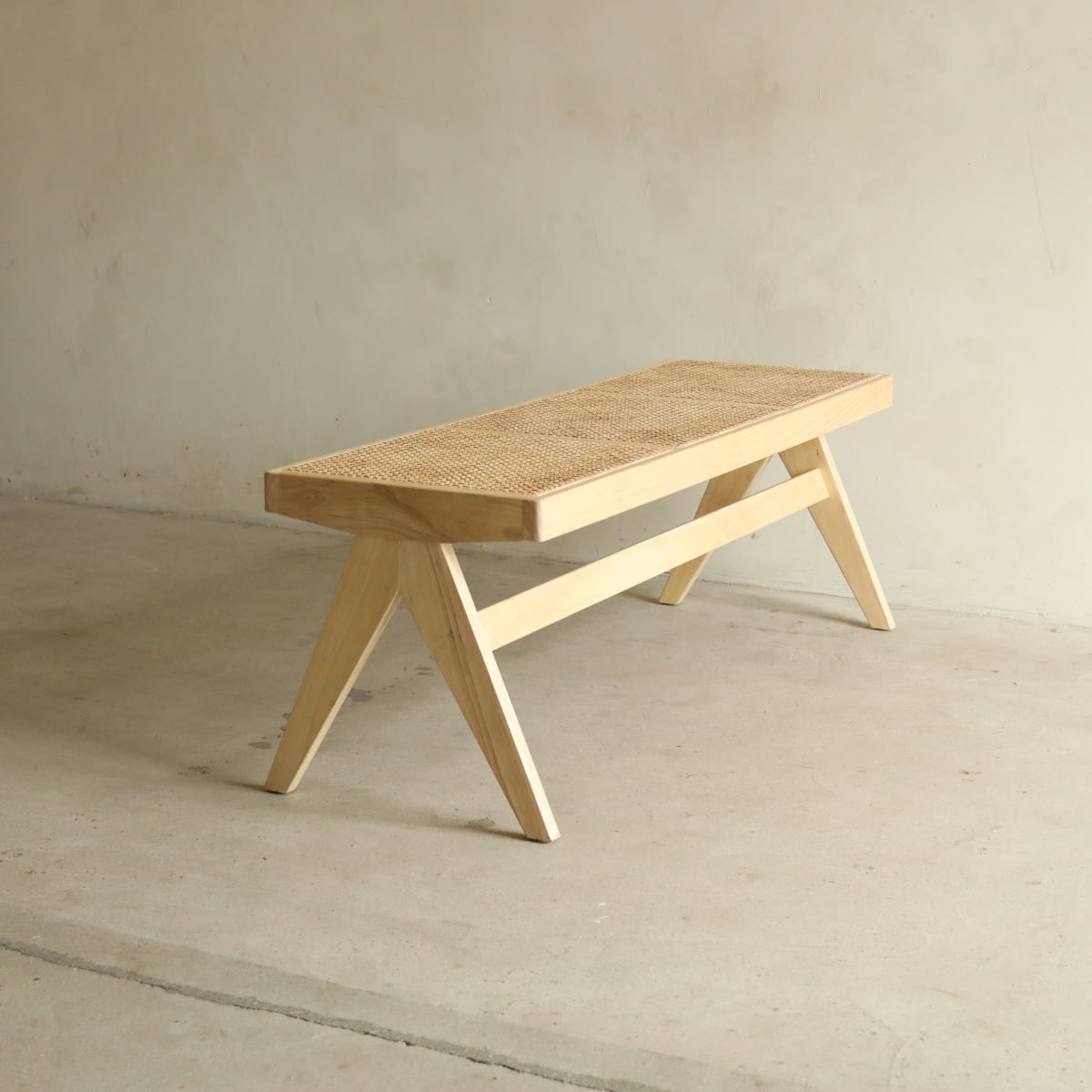 The Rivini Bench, crafted from teak wood with angled legs and a woven rattan seat in Scandinavian style, sits on a concrete floor against a plain, light-colored wall.