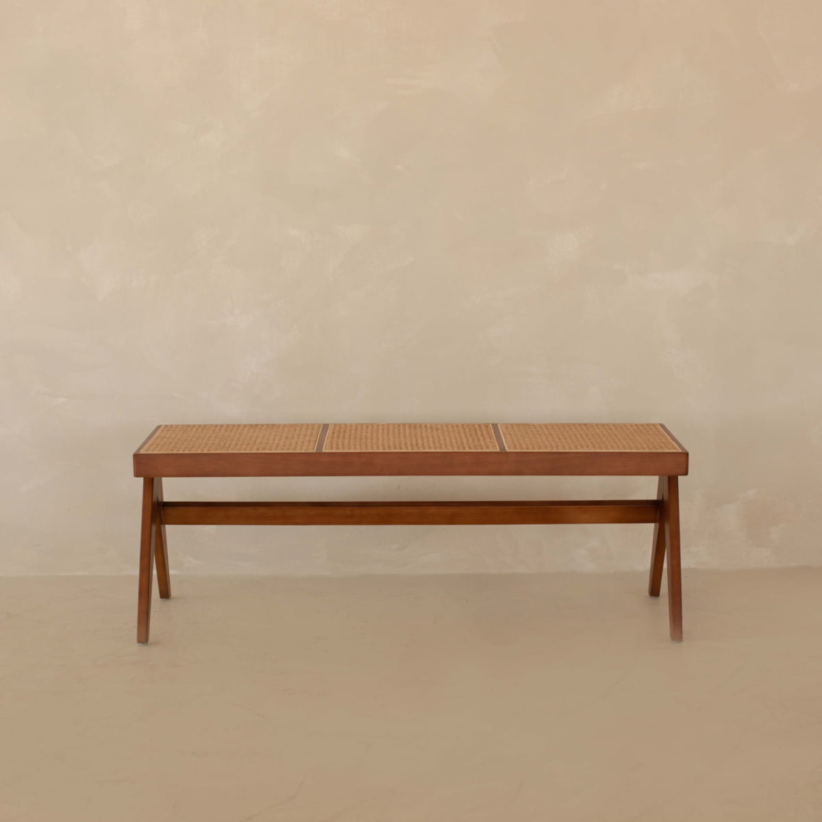 The Rivini Bench, crafted from teak wood with cane detailing, sits against a plain beige wall on a matching floor, showcasing understated Scandinavian style.