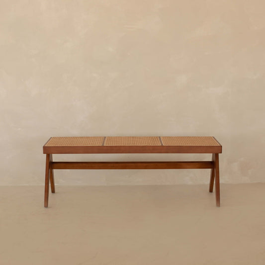 The Rivini Bench, crafted from teak wood with cane detailing, sits against a plain beige wall on a matching floor, showcasing understated Scandinavian style.