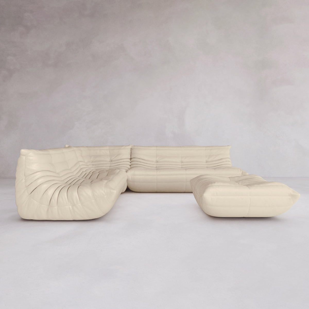 The Rolo Lounge Four Piece Sofa, cream-colored with a tufted plush design, sits on a light gray floor against a textured pale wall, showcasing its modular system for a minimalist and contemporary interior.