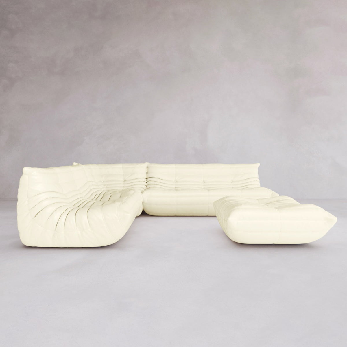 The Rolo Lounge Four Piece Sofa, featuring plush tufted upholstery and a modular design in cream, sits in a minimal light-gray room with bare walls and floor.