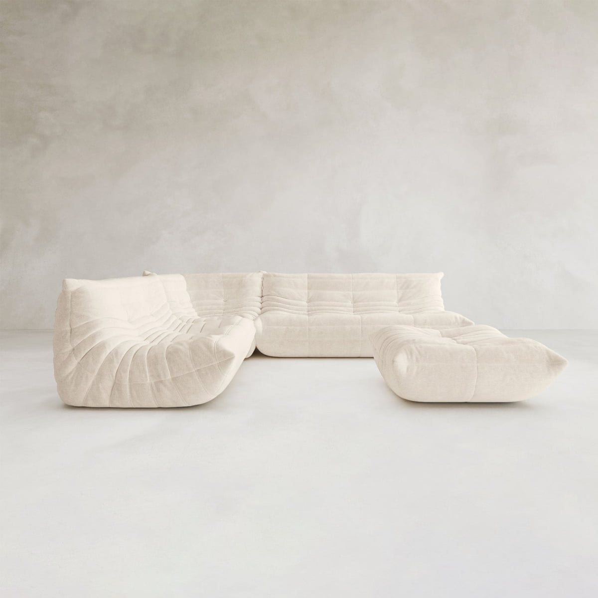 The Rolo Lounge Four Piece Sofa—a modern, modular cream sectional with deep cushions and tufted details—anchors a minimalist room with light gray walls and a smooth, light floor, evoking subtle mid century design.