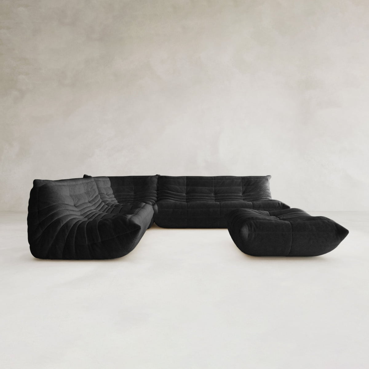 The Rolo Lounge Four Piece Sofa—a modern black modular sectional with deep, tufted cushions inspired by mid-century design—rests on a light floor against a neutral, textured wall.