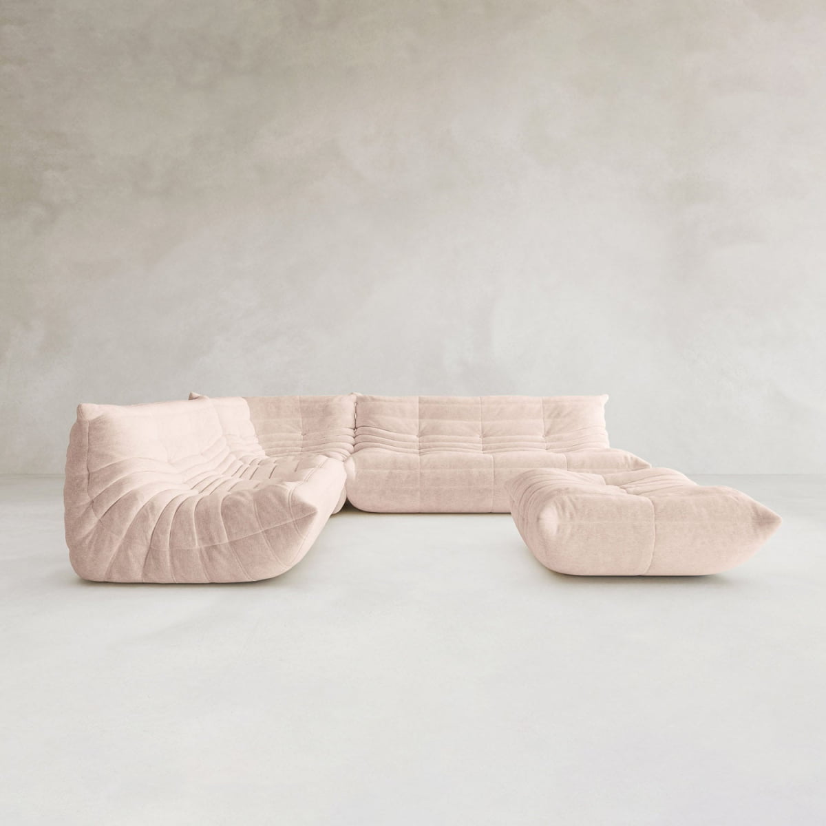 The Rolo Lounge Four Piece Sofa, a modern light beige sectional with a curved tufted design and mid-century influence, is featured in a minimalist room with pale walls and a smooth, light floor.