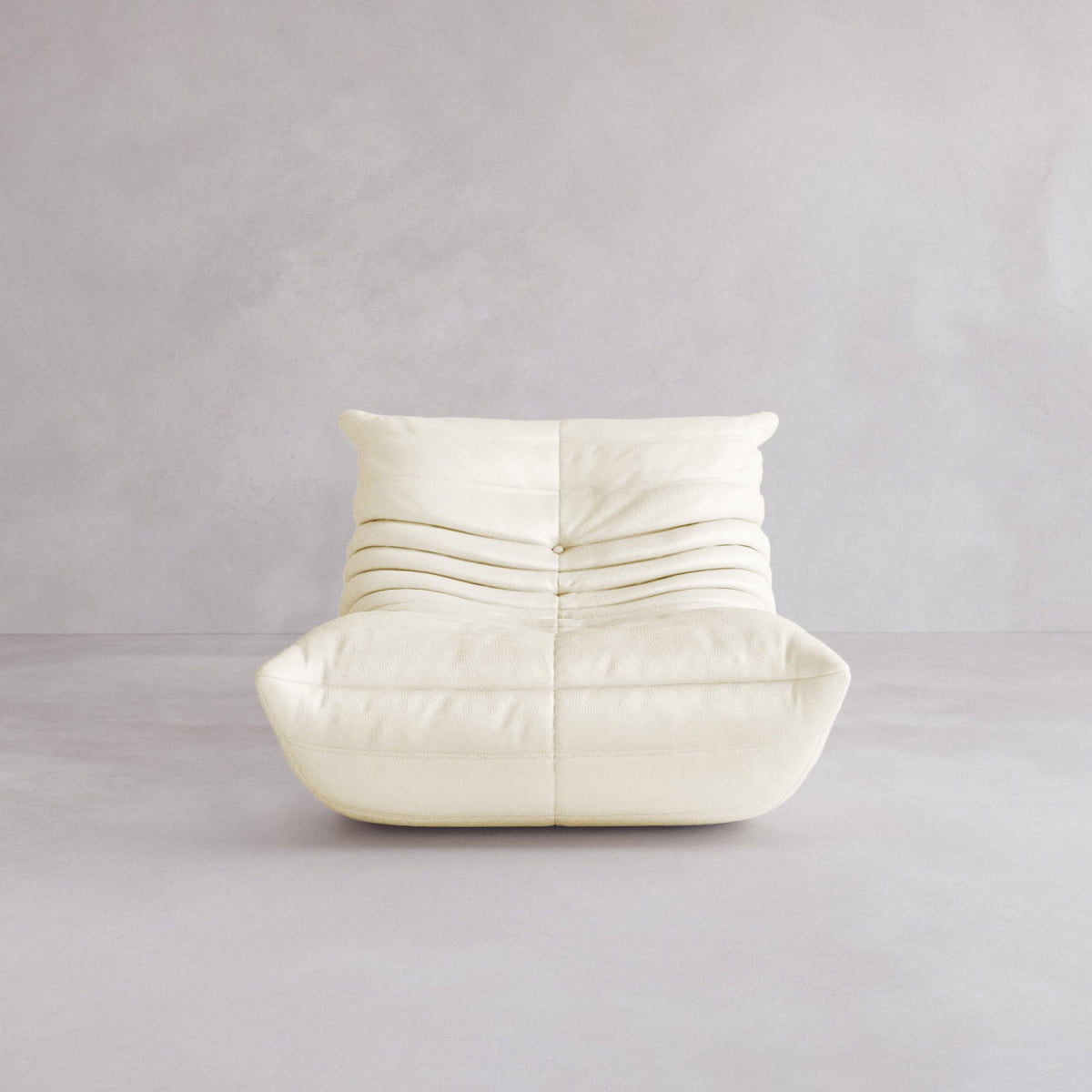 The Rolo Lounge Sofa Chair with Ottoman features a cream color, armless design, low wide seat, and horizontal ruched upholstery for a mid-century vibe, shown against a plain light gray background.