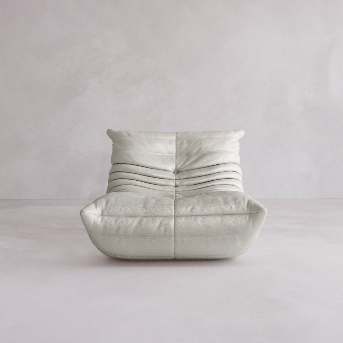 The Rolo Lounge Sofa Chair with Ottoman features soft light gray leather upholstery and horizontal pleats, offering a modern 70s-inspired design. It sits on a smooth pale floor against a light textured wall.