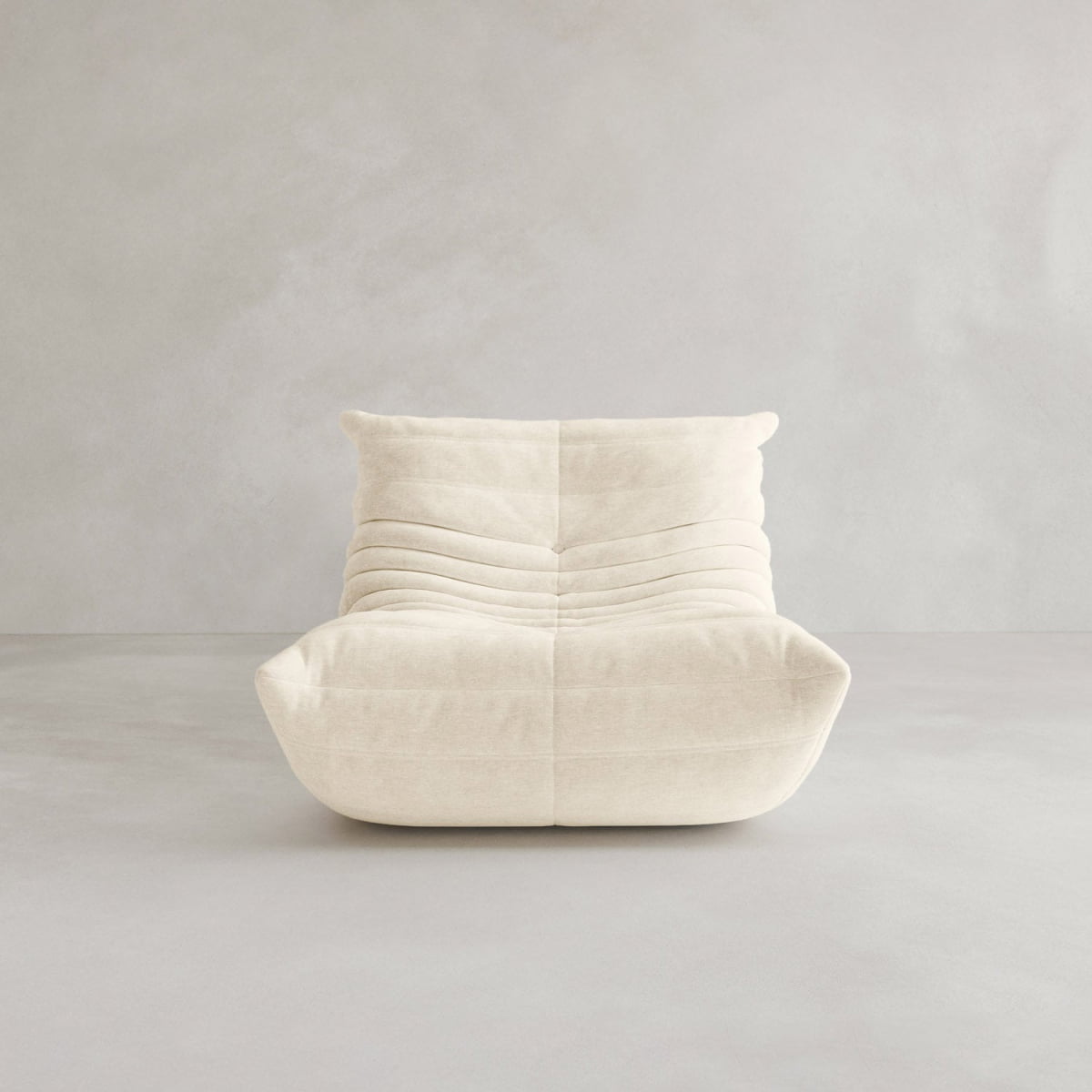 The Rolo Lounge Sofa Chair with Ottoman features modern, armless design, creamy beige upholstery with horizontal tufted folds, and a low mid-century profile, shown on a light gray floor against a matching gray wall.