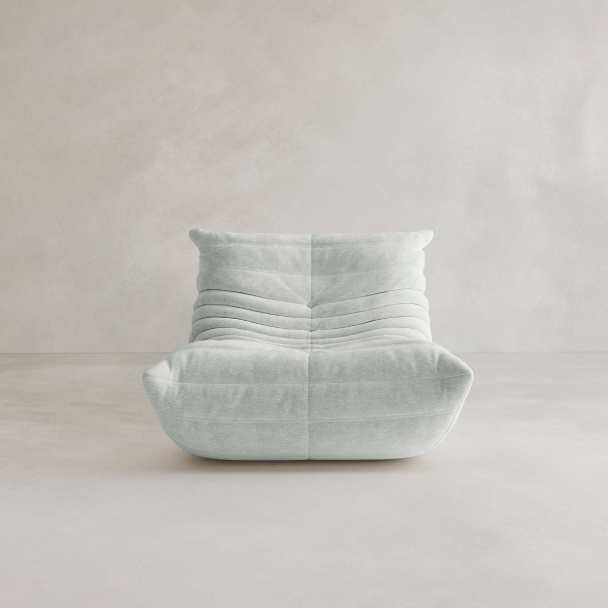 The Rolo Lounge Sofa Chair with Ottoman, featuring a modern light gray 70s-inspired design and plush gathered upholstery, sits on a pale floor against a neutral textured wall.