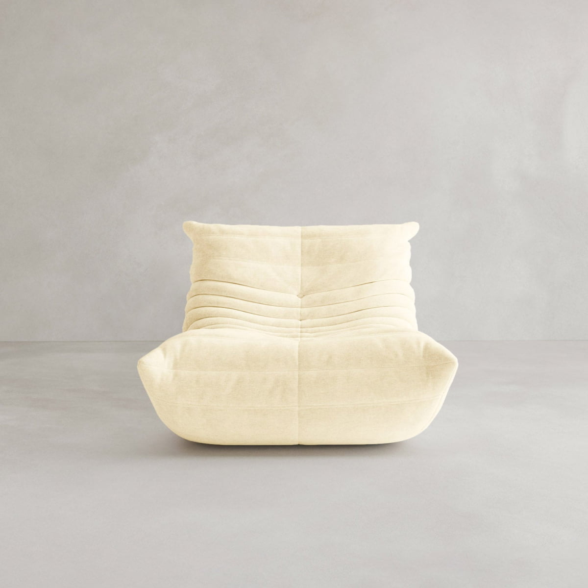 The Rolo Lounge Sofa Chair with Ottoman, inspired by 70s design, features soft light beige upholstery, plush horizontal tufting, and a low-profile boxy silhouette. It sits against a plain light gray wall and floor background.