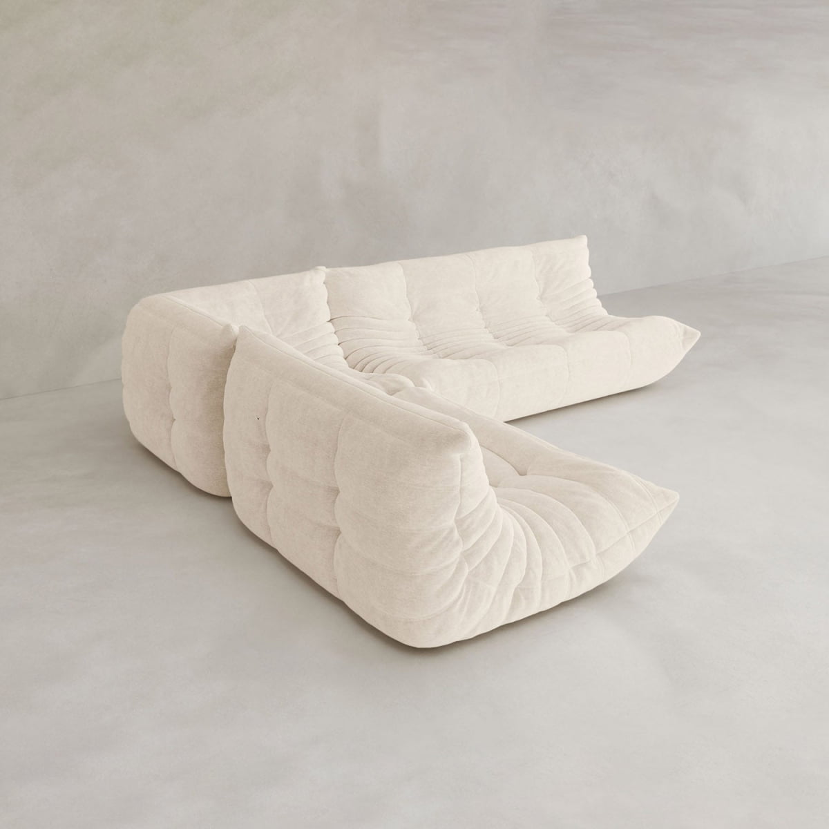 The Rolo Lounge Three Piece Sofa, a modern cream L-shaped sectional with deep tufted cushions, anchors the minimal light-toned room. Its modular design offers versatile layouts, while smooth walls and floors complete the mid-century inspired space.