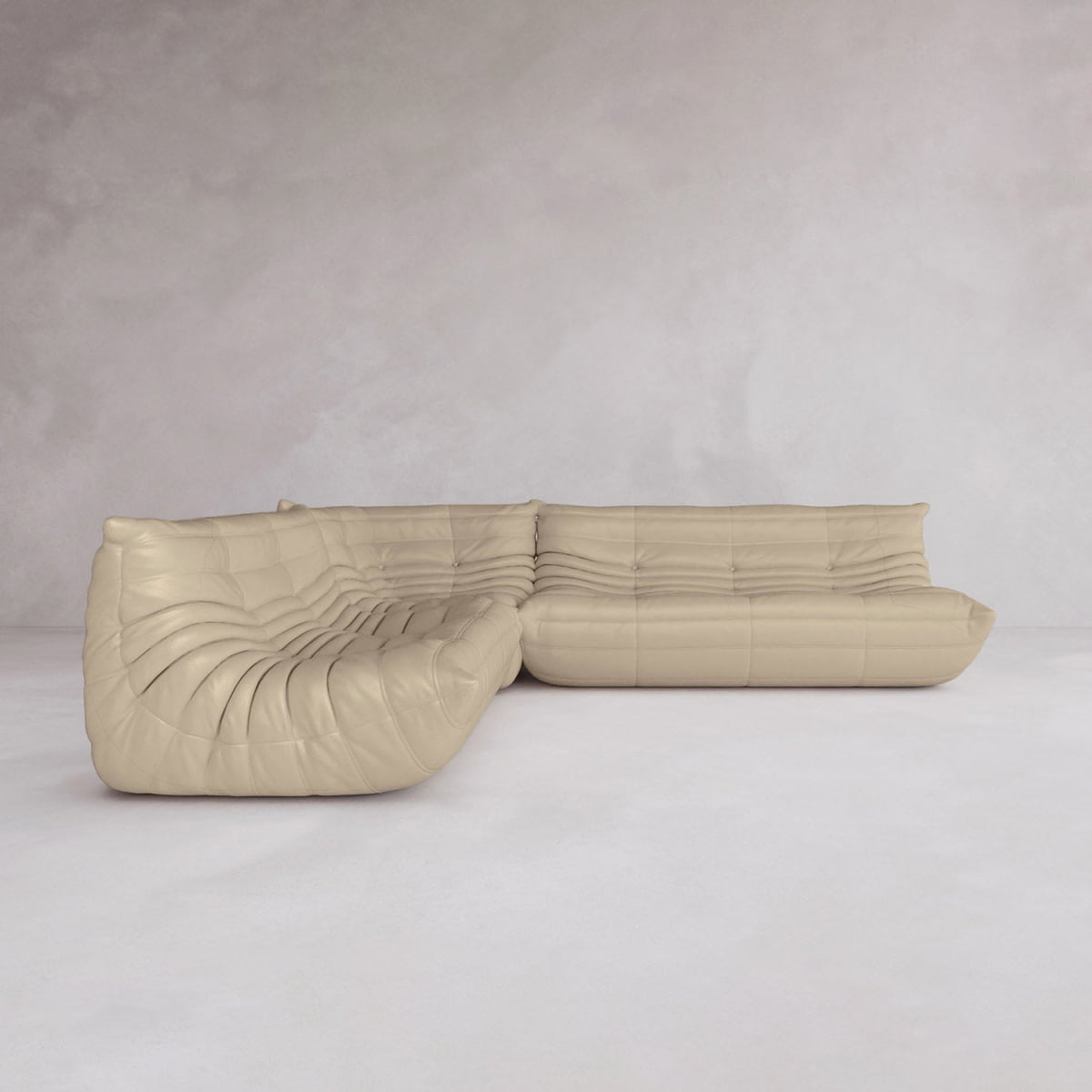 The Rolo Lounge Three Piece Sofa, a curved beige leather sectional with tufted plush cushions and modular design, is featured in a minimalist light gray room with smooth walls and flooring.
