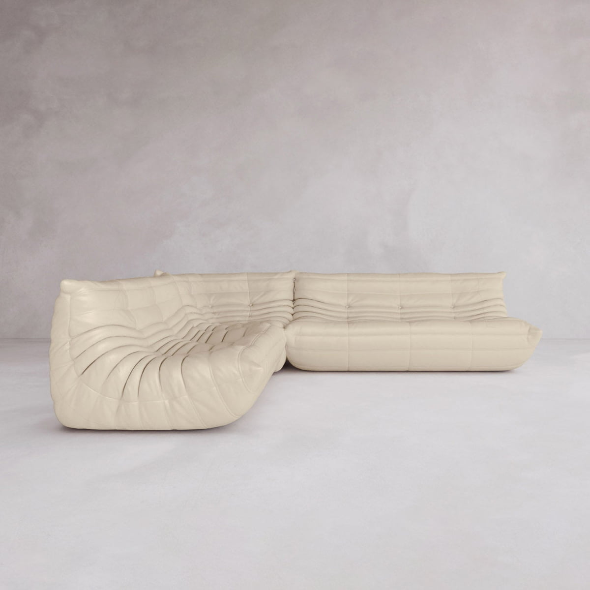 The Rolo Lounge Three Piece Sofa, a cream-colored modern sectional with a curved design and tufted upholstery inspired by mid-century style, sits in an empty room with light gray walls and floors.
