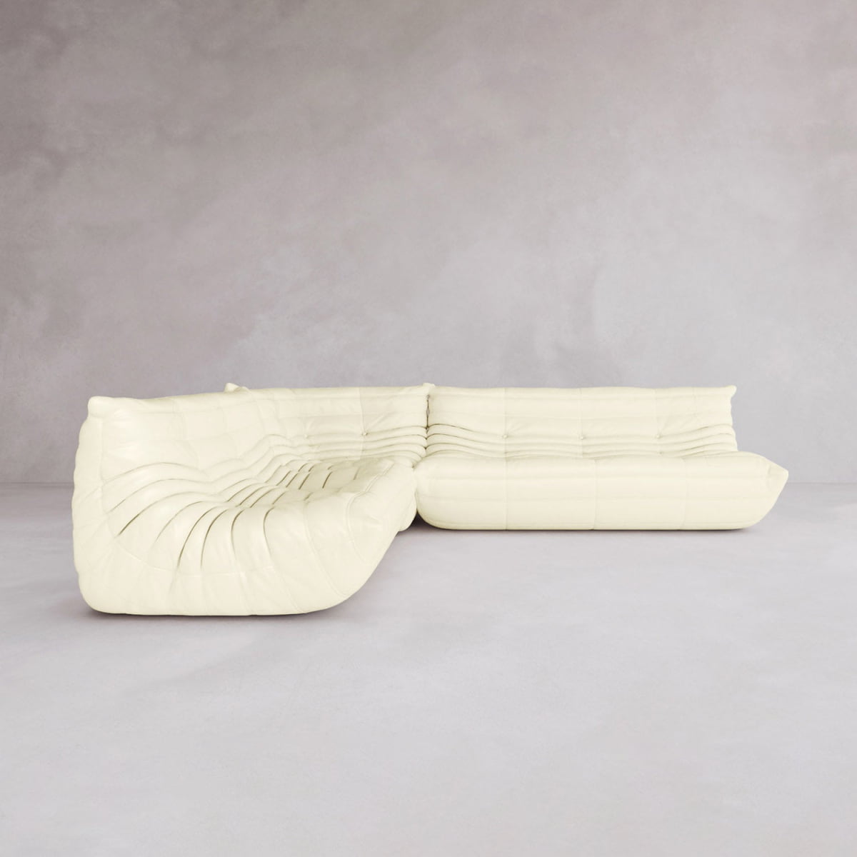 The Rolo Lounge Three Piece Sofa, a modern cream-colored sectional with curved mid-century design and plush tufted upholstery, sits on a light gray floor against a plain, light gray wall.