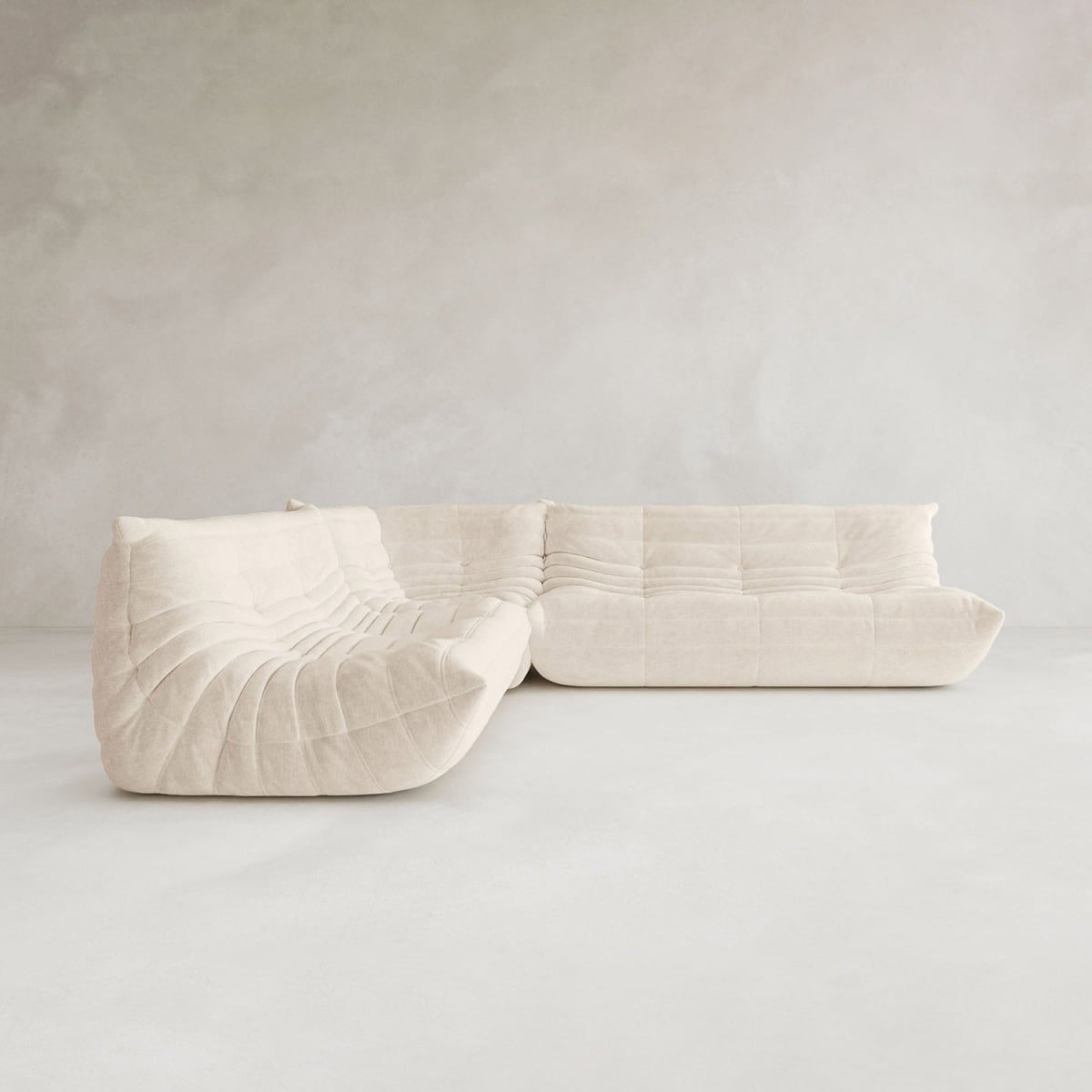 The Rolo Lounge Three Piece Sofa, a modular beige sectional with a tufted, plush look and modern design, is featured in a minimalistic, light-toned room with plain textured walls and floors.
