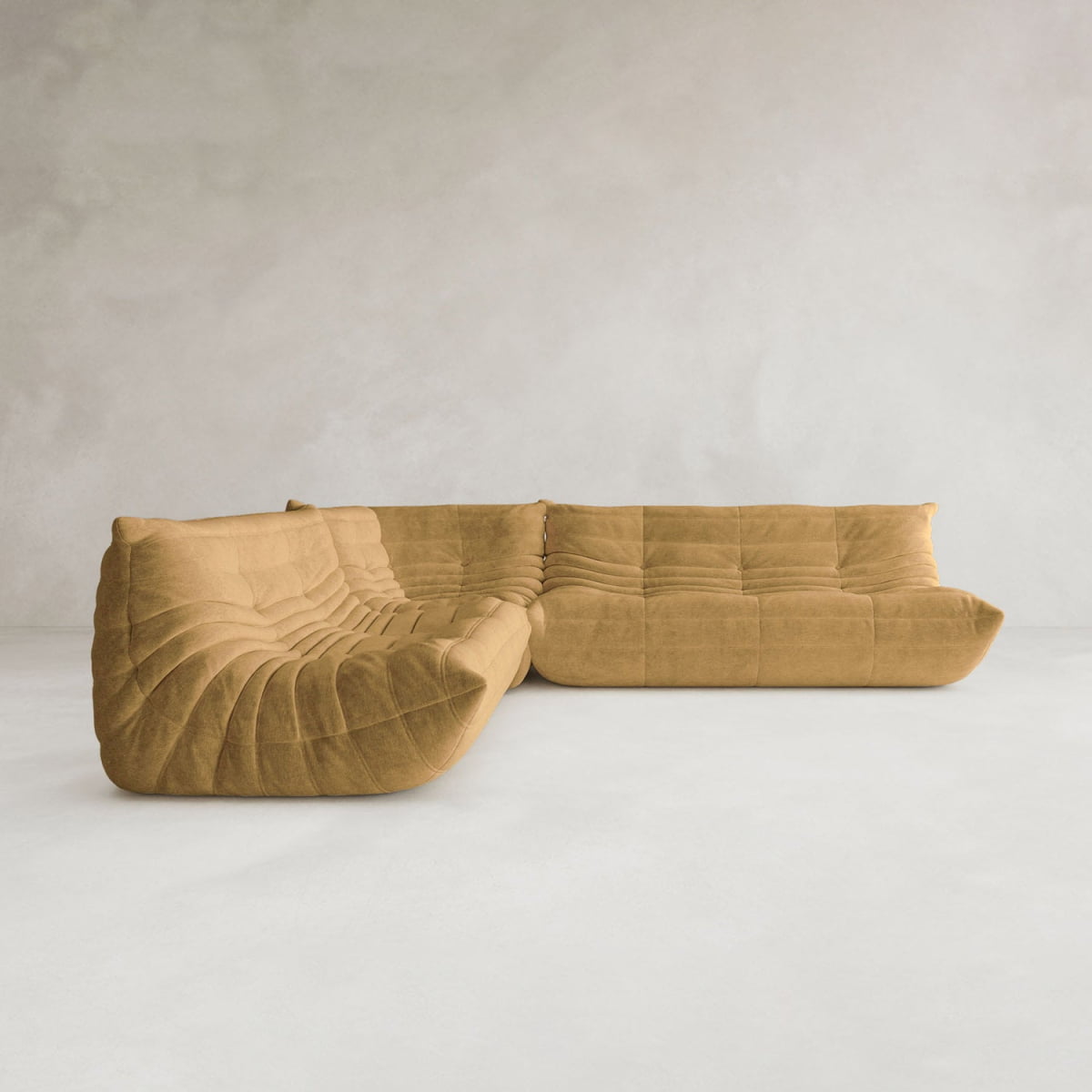 The Rolo Lounge Three Piece Sofa, a modern and modular tan sectional with a quilted plush texture and mid-century style, is placed against a plain light gray textured wall and floor in a minimalist setting.