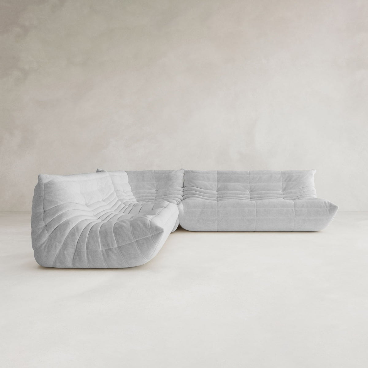 The Rolo Lounge Three Piece Sofa, featuring modern light gray tufted upholstery, sits in a minimalist room with pale walls and floors, offering versatile modular seating inspired by mid-century design.