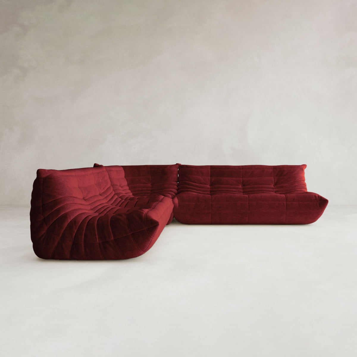 The Rolo Lounge Three Piece Sofa, a modern deep red velvet modular sectional with tufted cushions and a low profile, sits in a minimalist room with light gray textured walls and a light floor, adding mid-century flair.