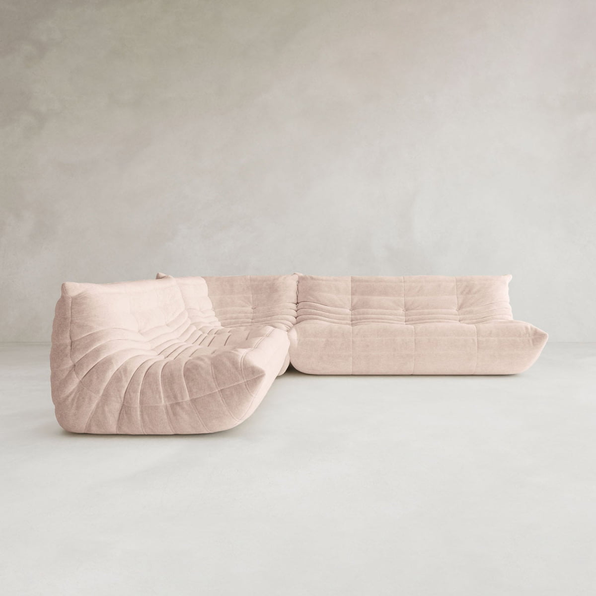 The Rolo Lounge Three Piece Sofa is a modern, light beige sectional with tufted plush design and curved lines, inspired by mid-century style, set against a plain, light-colored wall and floor.