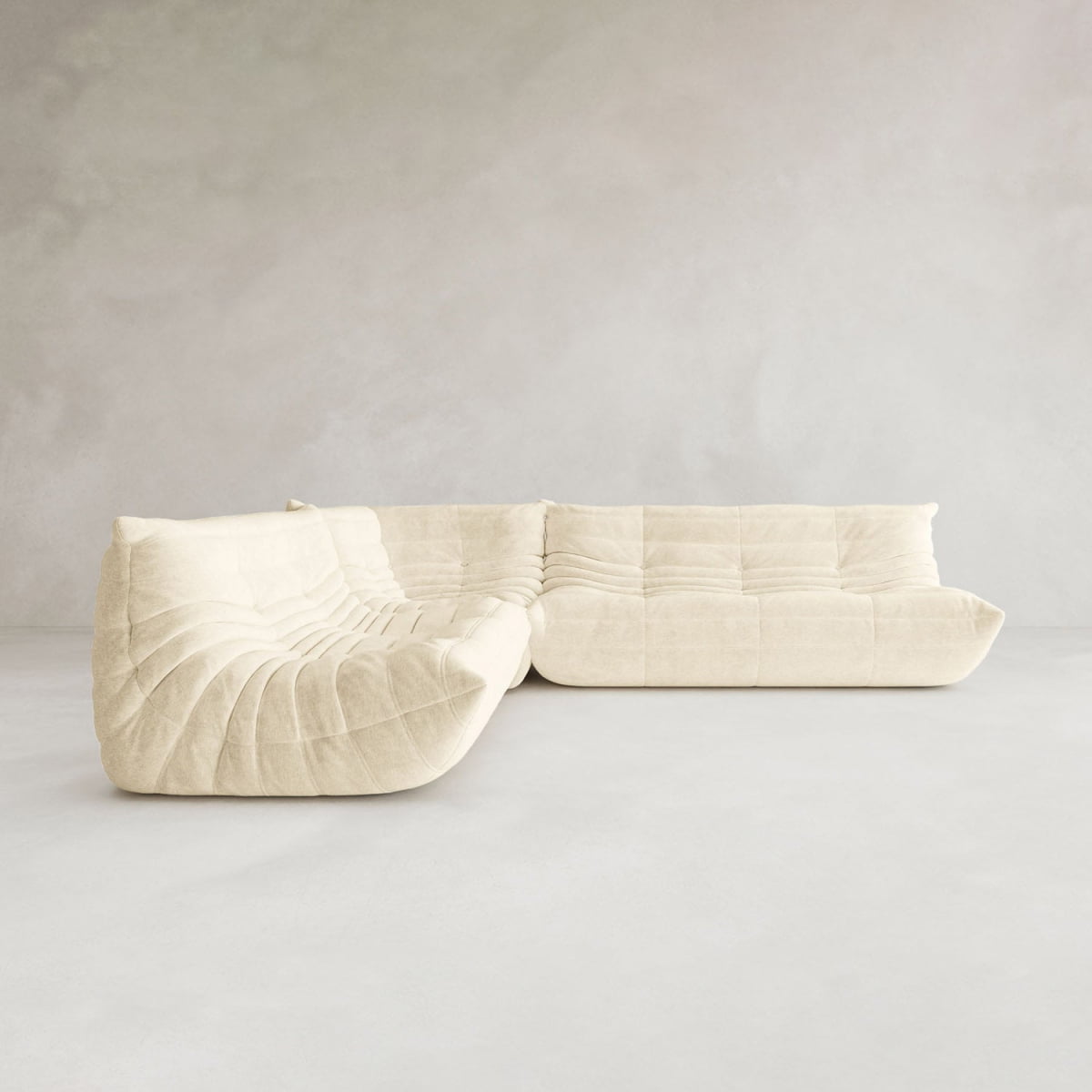 The Rolo Lounge Three Piece Sofa, a modern cream-colored modular sectional with soft tufted upholstery, sits in a minimalist room with light gray walls and a smooth floor, highlighting its contemporary design.