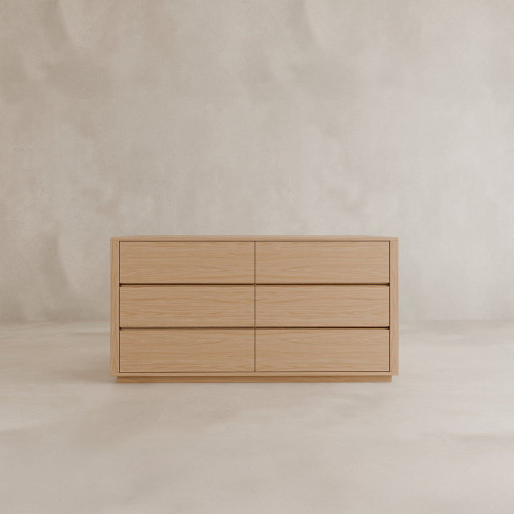 The Sandringham Dresser, featuring six spacious drawers, sits against a plain, light wall on a matching floor, bringing timeless minimalist elegance to the room.