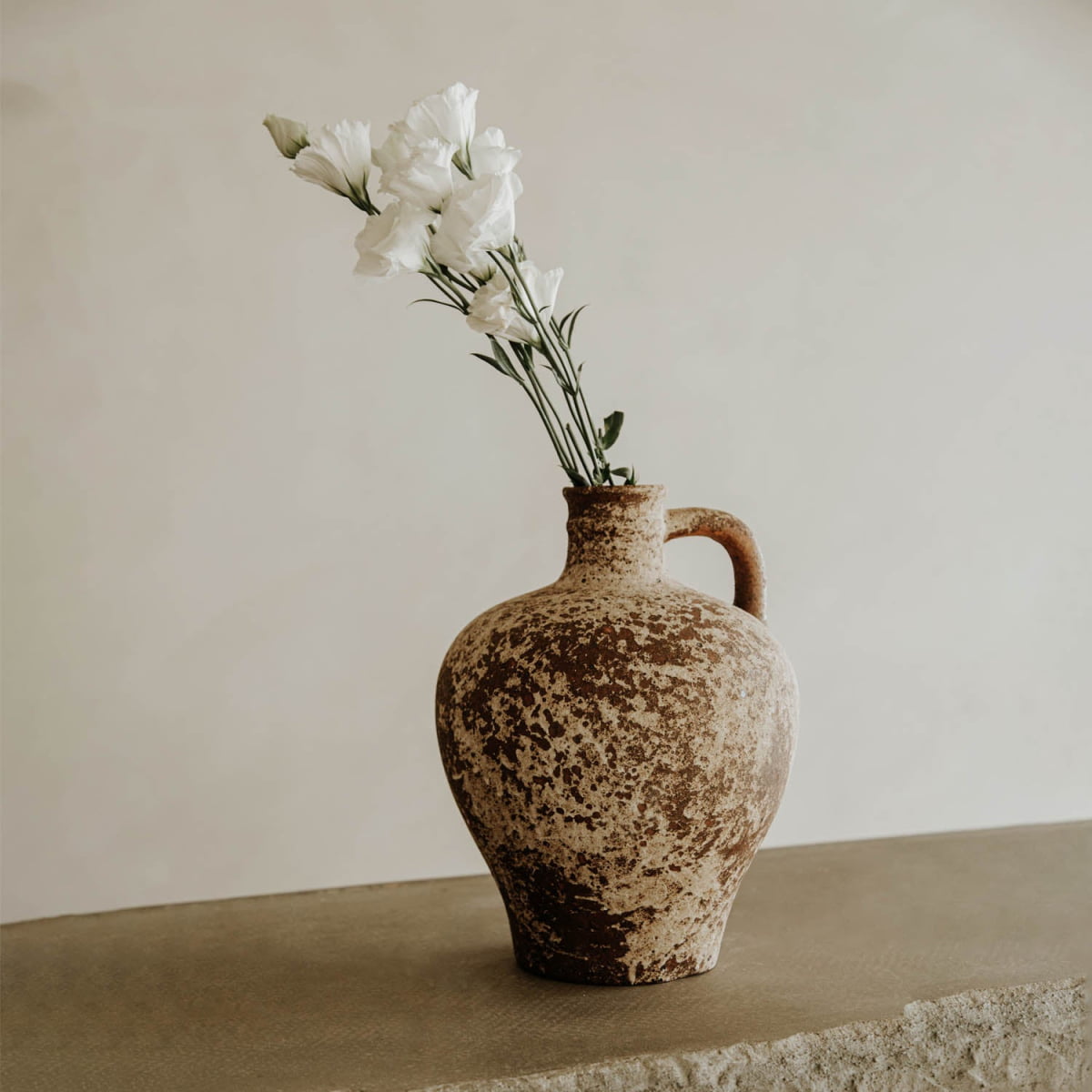 The Sato, a rustic textured ceramic jug inspired by artisan pottery, displays a small bouquet of white flowers against a neutral background on a stone surface.
