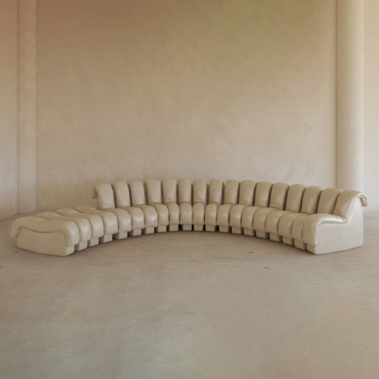 The Serpenti Sofa 15 Piece With Ottoman Rental is a large, curved beige leather modular system with tufted cushions and addable armchair elements for flexible seating, set against a plain light wall in a minimalist room.