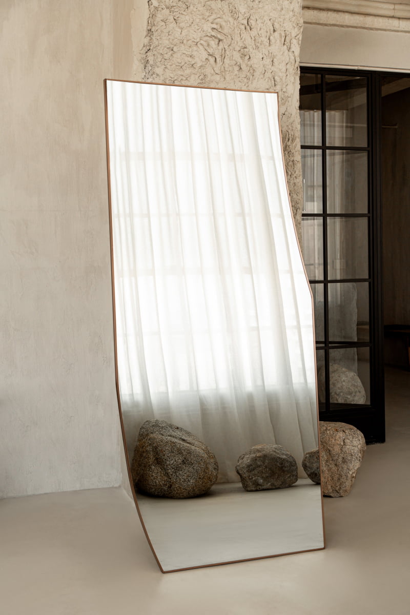 The Shard Mirror L reflects sheer white curtains and two large rocks in a minimalist, neutral-toned room with textured walls and a black-framed glass door, capturing modern elegance.